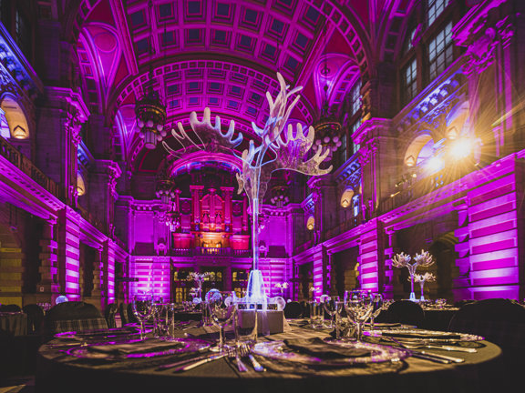 An internal view of the Kelvingrove Main Hall decorated for an event. The view from the ground floor shows a cavernous, vaulted, 3 storey space with carved stone walls, lit with pink and golden lights.  A huge pipe organ is visible on the first storey balcony at the far end of the room. The foreground is dominated by a round table, decorated with a black table cloth, glasses and silverware. A transparent, acrylic, lit centrepiece - resembling antlers or a tree branch, catches the glow of the spotlights.
