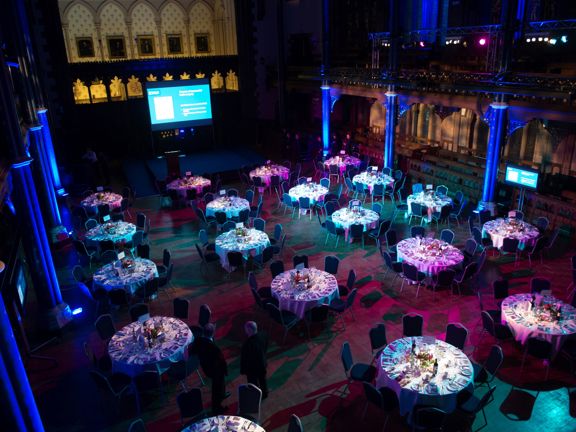 The UoG's Bute Hall arranged for a private dinner event, looking down from a first floor balcony. A large hall with wooden floors is lit with pink, blue and turquoise spotlights. The edge of the room is framed with carved stone pillars that support the balcony, they are lit blue. Dozens of round tables with white table cloths are laid with silverware, cut flowers and crockery; each table is surrounded by 8 chairs. A small temporary stage and a large projection screen are visible at the far end of the room.