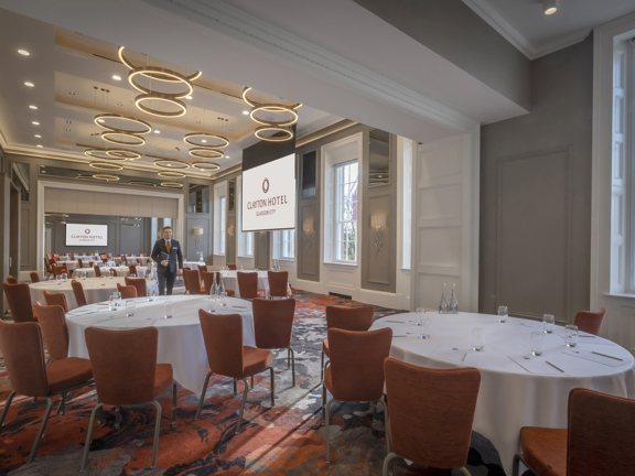 An interior view of a room in the Clayton Hotel. The room is arranged with orange chairs surrounding multiple round tables with white table cloths. The room is decorated with bold patterned carpets in shades of orange and grey and modern circular over-head lights, contrasting with the original wooden panelling, sash windows and decorative plasterwork details. A large television is visible on the far wall, while a projection screen hangs in the centre of the room, both are showing the Clayton Hotel branding.