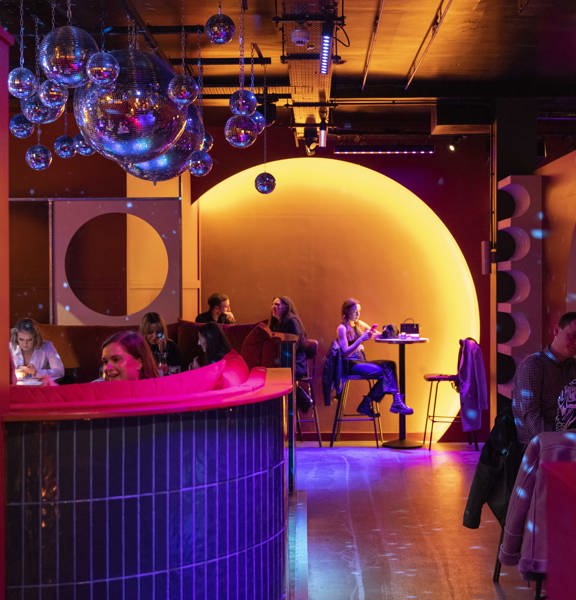 An interior view from August House shows a vibrant bar scene with a mix of high and low seats and tables. A large accent wall at the end of the room has a large circle set into the wall and lit orange. Other arched and round details cut out of walls repeat the motif. Disco balls hang from the ceiling, clustered together, throwing specs of blue light over the otherwise orange lit space. The floor looks smooth and polished.