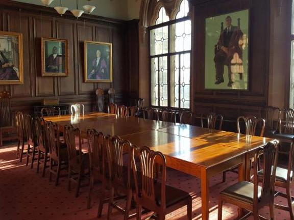 Old wood panelled room with pictures of people on the walls hung in ornate gold frames. Large wooden boardroom table sits in the middle of the room surrounded by wooden chairs 