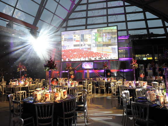An interior view of the Glasgow Science Centre decorated for an event.  The roof is windowed, though nothing can be seen outside as it is dusk. The room is filled with round table with black cloths, gold chairs surround them. The space is dark, spotlights, a large projected screen and lit centre pieces on the tables light the room. The tables are decorated with menus, silverware, vases of flowers and lights. A small stage and scaffold are just visible, there is a lectern and television screen visible on it.
