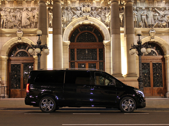 A large, black, shiny people-carrier is parked outside of a white stone building with fine figurative carvings and pillars. Large wooden doors and 2 ornate lampposts frame the picture symetrically. 