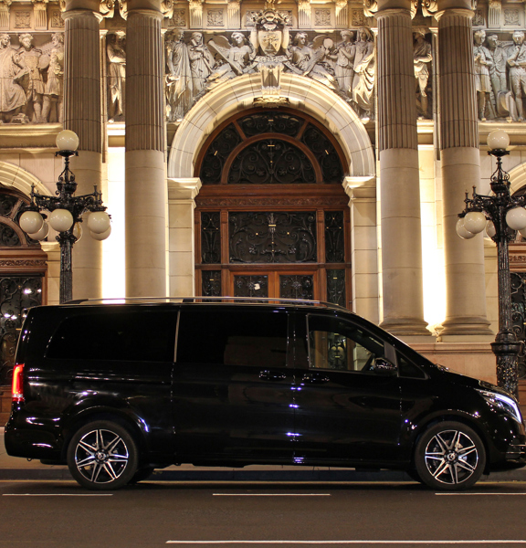 A large, black, shiny people-carrier is parked outside of a white stone building with fine figurative carvings and pillars. Large wooden doors and 2 ornate lampposts frame the picture symetrically. 