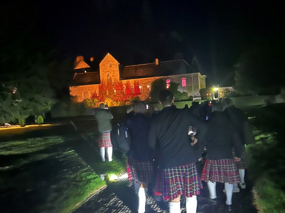 Large stone period building set in the countryside amongst trees. A large group of people wearing kilts walk on cobbled path towards the house at night.