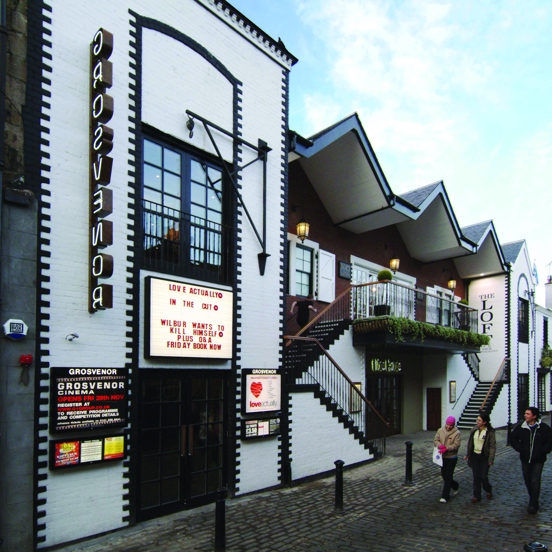 An exterior view of the Grosvenor cafe and cinema shows a white rendered building with black-painted brick details, it boasts features like first-storey windows and pulleys from the buildings original purpose. An entrance on the left hand side has a lit sign reading "Grosvenor," vertically, there is also a lit board with film listings and further posters below. 2 symmetrical staircases lead to a balcony, beside the stairs there is text reading, "The Loft." The building is on a cobbled lane with bollards.