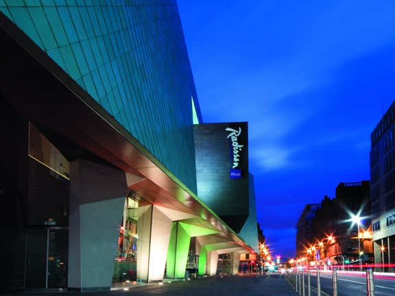 An exterior image of the Radisson Blu hotel. Shows a modern, angular building, The ground floor has glass walls, punctuated by angular, white, concrete pillars; the photo is taken at night and the pillars are brightly lit. The visible facade of the building is covered in diamond-shaped, turquoise tiles. A large, grey prism juts out of the first floor bearing the Radisson Blu logo. The hotel is on a wide, paved pavement, bollards define the line between the pavement and the streetlight lit road beyond.