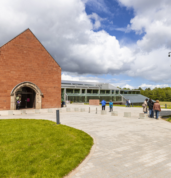 An external view of the Burrell Collection. A paved walkway leads up to the red sandstone entrance of the museum. A large glass building and green lawn is visible against trees and a cloudy sky.