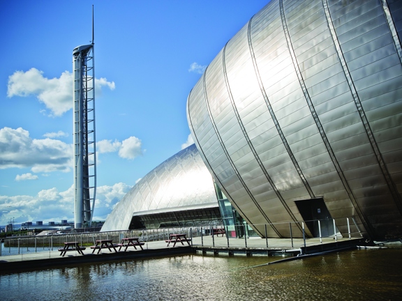 An exterior image of the Glasgow Science Centre shows the end of 2 modern, bulbous buildings; their surfaces are made up of silver plating. In the foreground there is a fenced off pool of water, a paved walkway leads over the water to picnic benches that sit at the base of the closest building, they are dwarfed by its scale. A third structure to the left is the Glasgow Tower, a tall, free-standing white and grey, tubular, observation tower. The sky is blue with large white clouds.