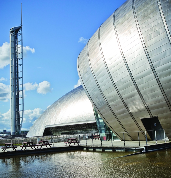 An exterior image of the Glasgow Science Centre shows the end of 2 modern, bulbous buildings; their surfaces are made up of silver plating. In the foreground there is a fenced off pool of water, a paved walkway leads over the water to picnic benches that sit at the base of the closest building, they are dwarfed by its scale. A third structure to the left is the Glasgow Tower, a tall, free-standing white and grey, tubular, observation tower. The sky is blue with large white clouds.