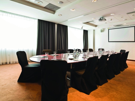 A photo of the interior of a Radisson Blu meeting room. A long, wooden, oval table sits in the centre of an auburn-carpeted room. he walls are white, and the long, far wall has 2 large floor to ceiling windows. Heavy fabric curtains are pulled back on each window, but mesh curtains stop us seeing out. 10 chairs covered with black cloth surround the table which has glass water bottles and stationary. A projector can be seen mounted on the ceiling and a projector screen and 2 speakers are mounted to the wall.