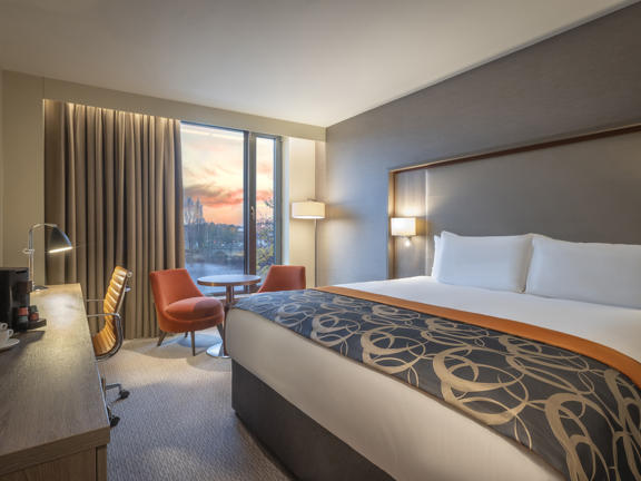 An interior shot of a bedroom at the Clayton Hotel. The room is decorated with a light coloured, patterned carpet and grey walls. There are 2 orange armchairs either side of a small round table, arranged in front of the floor to ceiling window. The bed is large and clean with a patterned blanket across the foot of it. A desk, desk chair, television and lamp are all visible on the left-hand side of the room.