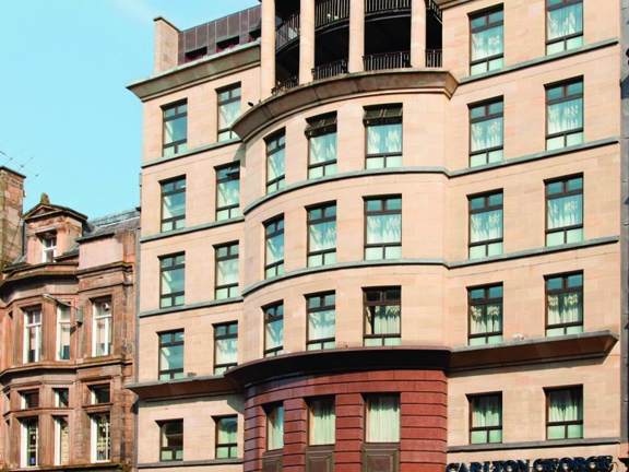 An external view of the Carlton George hotels, a multi-storey stone building. The top 2 floors have balconies and 4 large pillars. At street level shops and bars are visible.