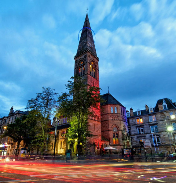 An exterior view of Oran Mor shows a Victorian sandstone church building on a street corner, with 2 large trees in its grounds. The image is taken at dusk with a long exposure so streaks of light and car headlights fill the road. The steeples windows are lit and the building is up-lit with red lights. Sandstone tenement buildings line the streets in either direction. The image is taken from low down so a large expanse of cloudy sky can be seen.