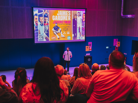 Person standing in front of a large audience in front of a large screen that reads:  James Gardner Journeyman