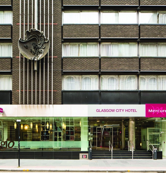 An exterior image of Mercure Hotel shows a 1960s building made of dark brick, 4 storeys are visible but it is possible to interpret that it is taller, the bedroom windows are broad. The pavement outside is wide and made up of paved stone. The ground floor has floor to ceiling windows, on the right a few, wide, black stairs lead up to the glass doorway, 3 silver handrails are visible. A silver and pink sign reads, "Glasgow City Hotel" and "Mercure". The facade is decorated with an abstract, metal artwork.