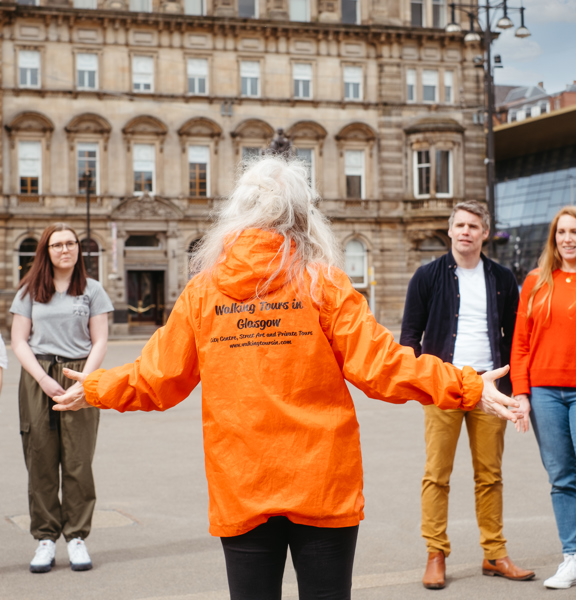 Four individuals, 3 women and one man, stand facing a tour guide with long white hair in a bright orange jacket. The tour guide has her back to us and her arms spread, expressively. Her jacket has black writing, obscured partially by her hair, it is possible to read "Walking Tours Glasgow". The group are stood on an expanse of paved ground. Tall sandstone buildings, a modern glass building, benches and a bronze statue on a plinth are visible behind them.