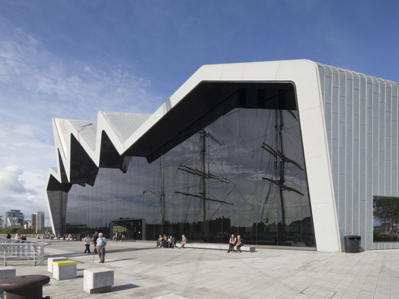 Sunny view of the modern, glass-fronted Riverside Museum, where the roofline reminiscent of a heartbeat on a monitor, with people enjoying the sunshine. The reflection of tall ship masts can be seen in the glass front of the museum